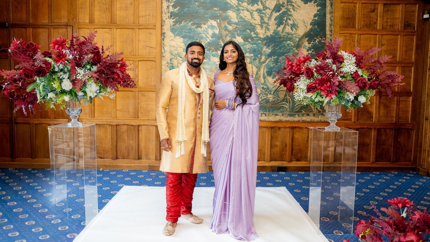 South Asian couple in gold sherwani and lavender saree posing at a floral decorated wedding venue.
