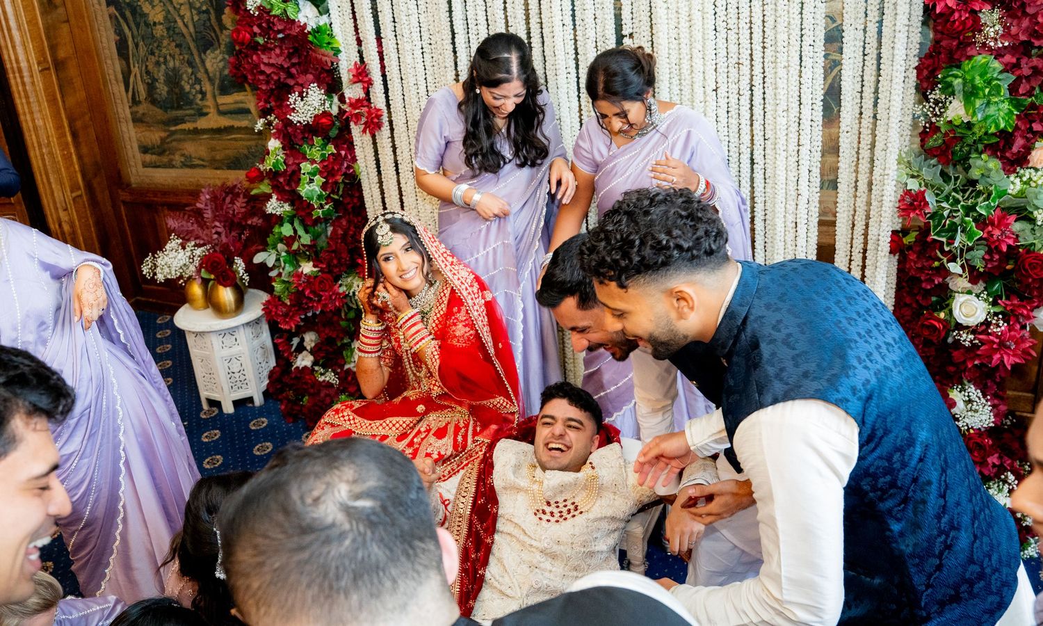 Joyful Indian wedding ceremony moment with bride in red lehenga surrounded by laughing family and floral decorations.
