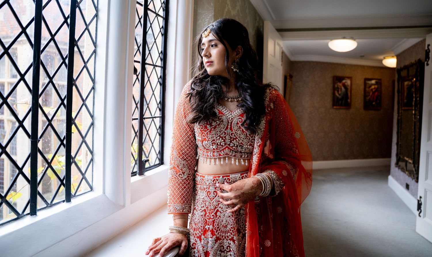 Indian bride in red lehenga gazes through latticed window in soft natural light.