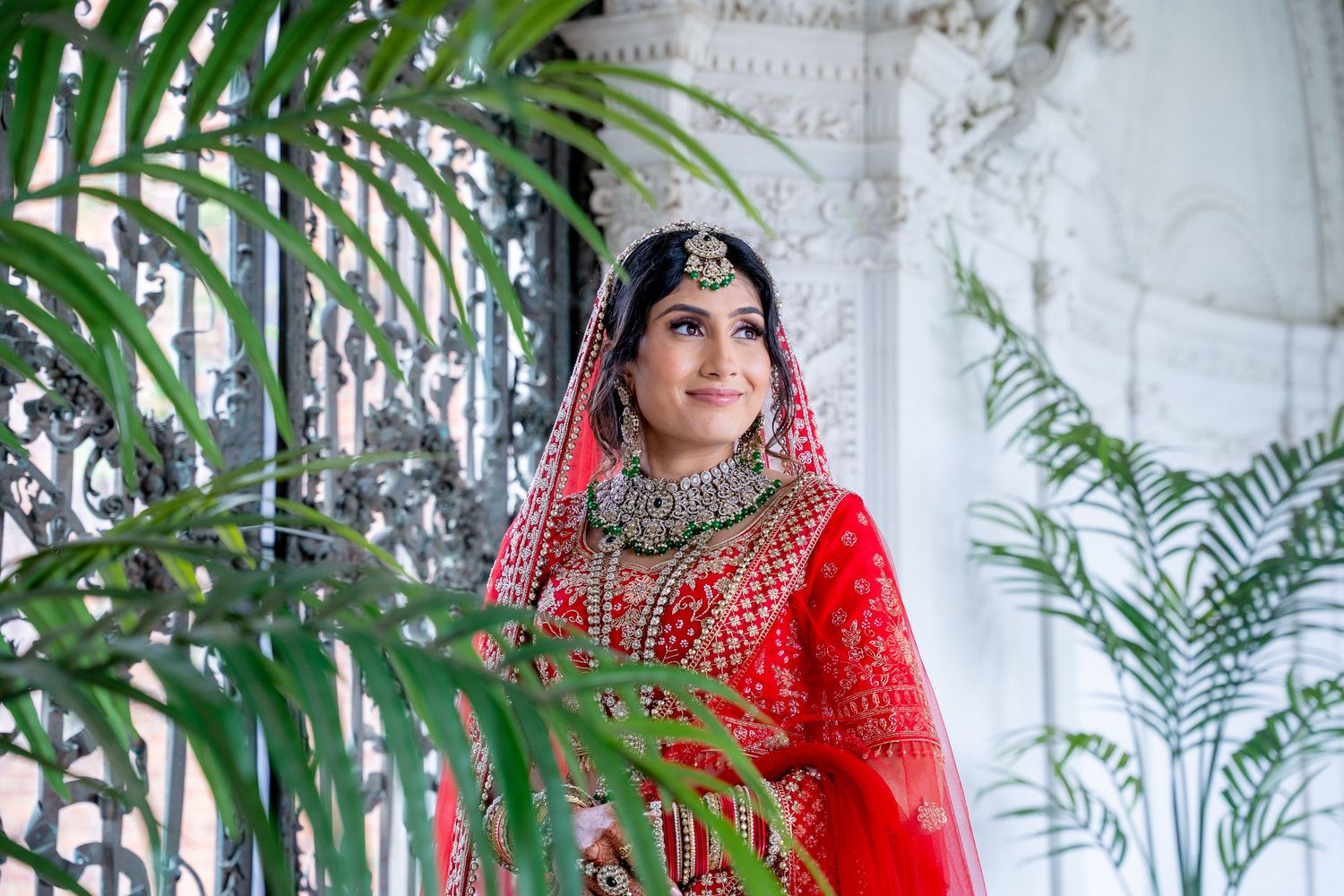 Beautiful Indian bride in red lehenga and ornate jewelry gazing upward near decorative ironwork and tropical plants.