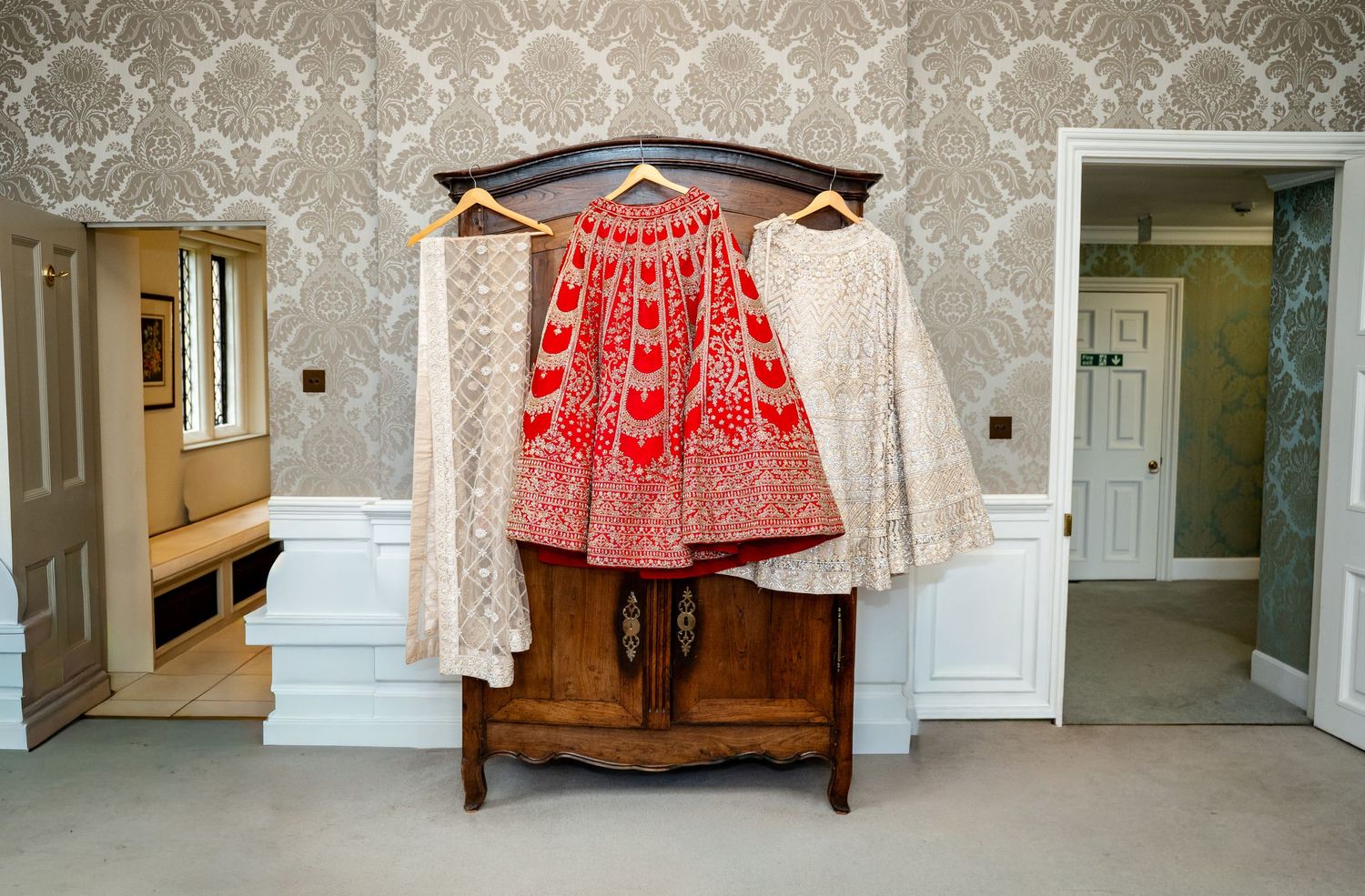 Red and white embroidered Indian bridal lehenga and dupatta hanging on wooden dresser in bedroom.