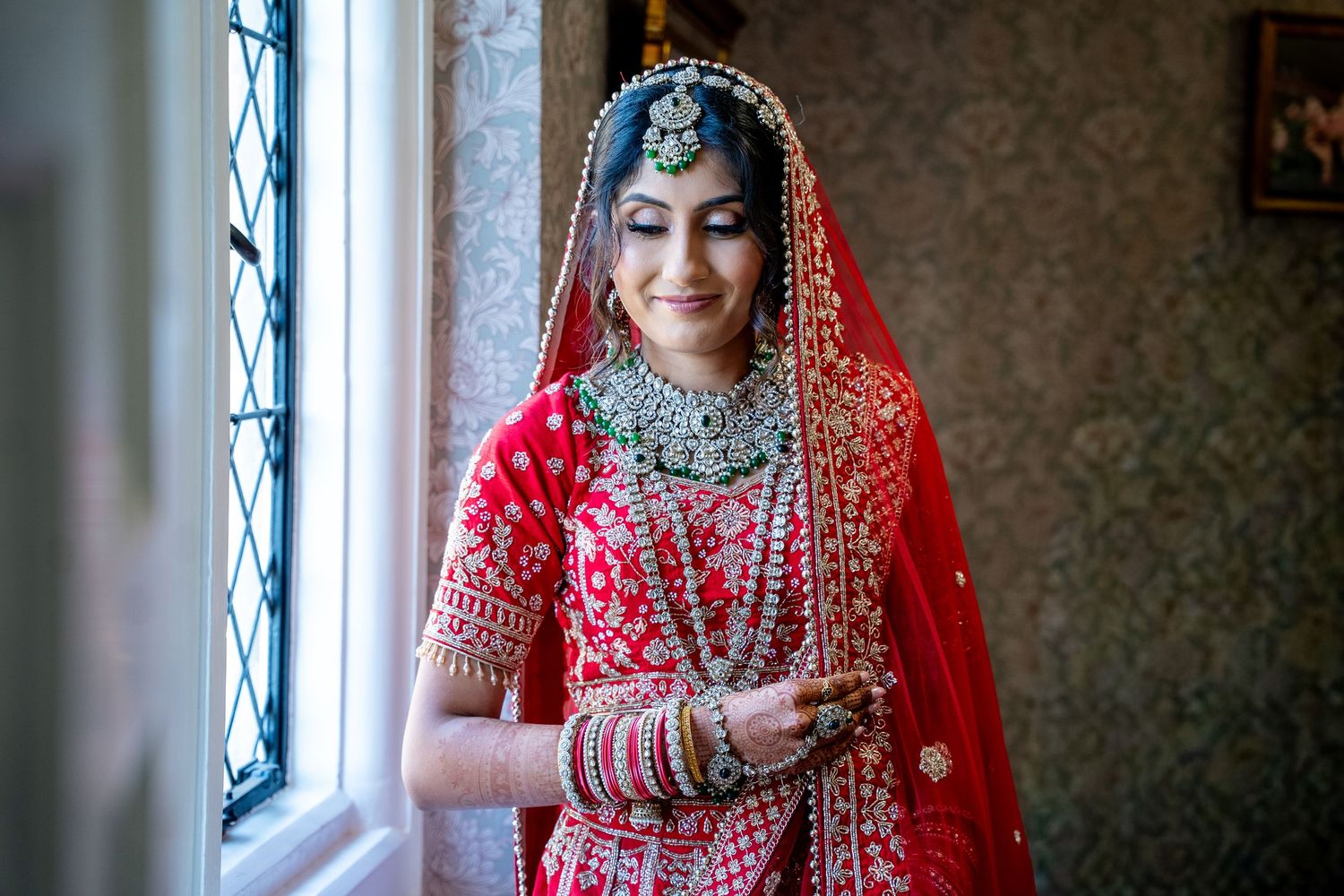Indian bride in red bridal lehenga and jewelry stands gracefully by a window in soft light.