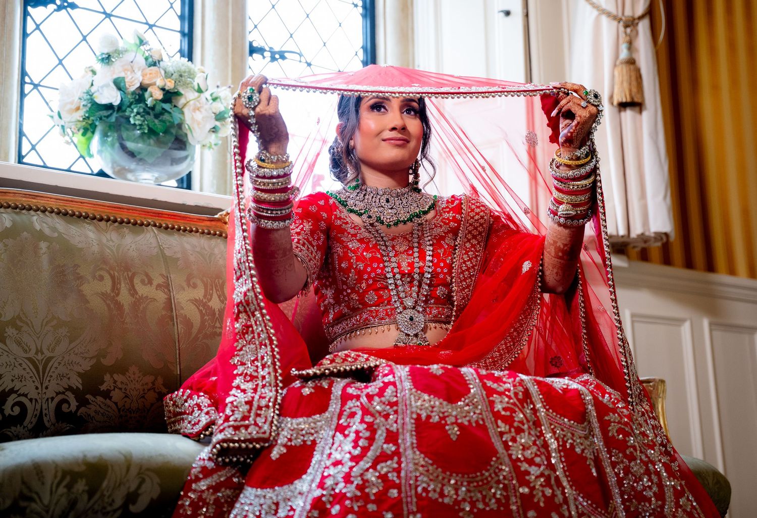 Indian bride in red lehenga holding dupatta overhead near window with floral bouquet.