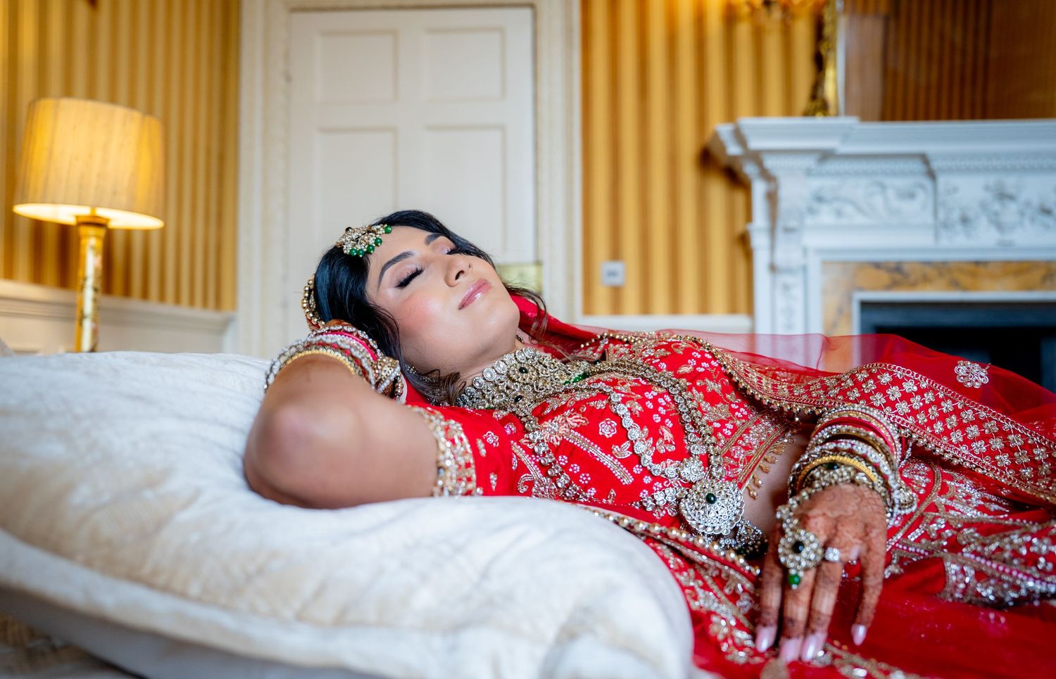 Indian bride in red bridal lehenga resting on white surface adorned with gold jewelry and bangles.