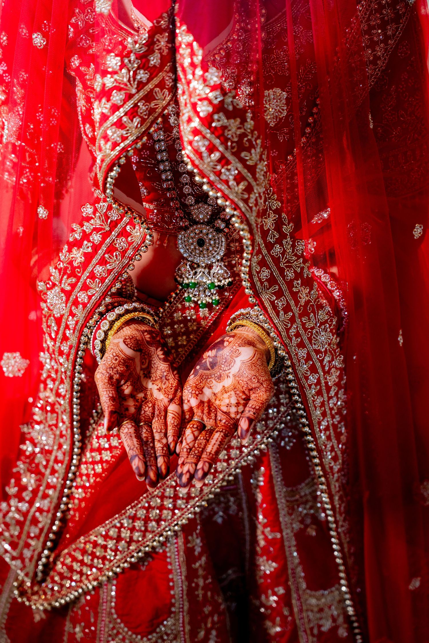 Close-up of Indian bride's henna-adorned hands holding embroidered red bridal lehenga with jewelry.
