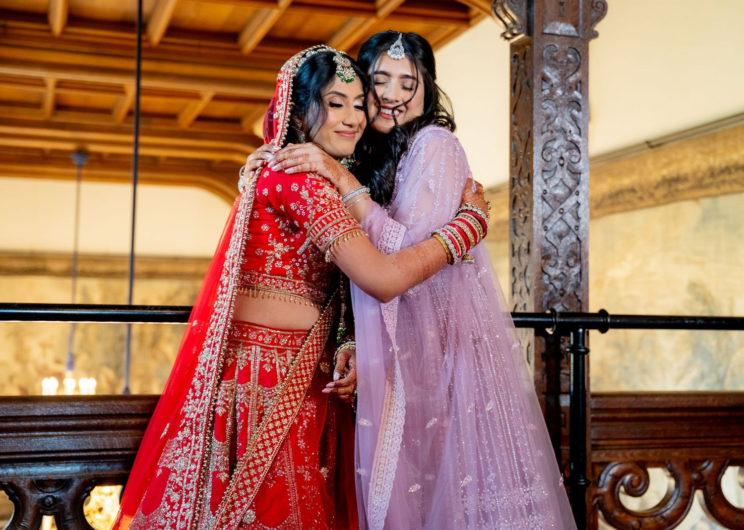 Indian bride in red lehenga shares emotional embrace with bridesmaid on ornate balcony.