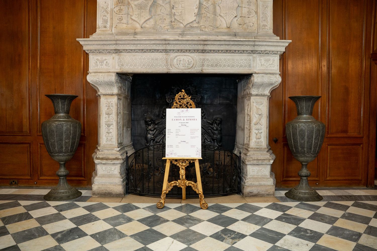 Elegant marble fireplace with ornate gold easel displaying a menu sign, flanked by two large decorative urns on checkered floor.