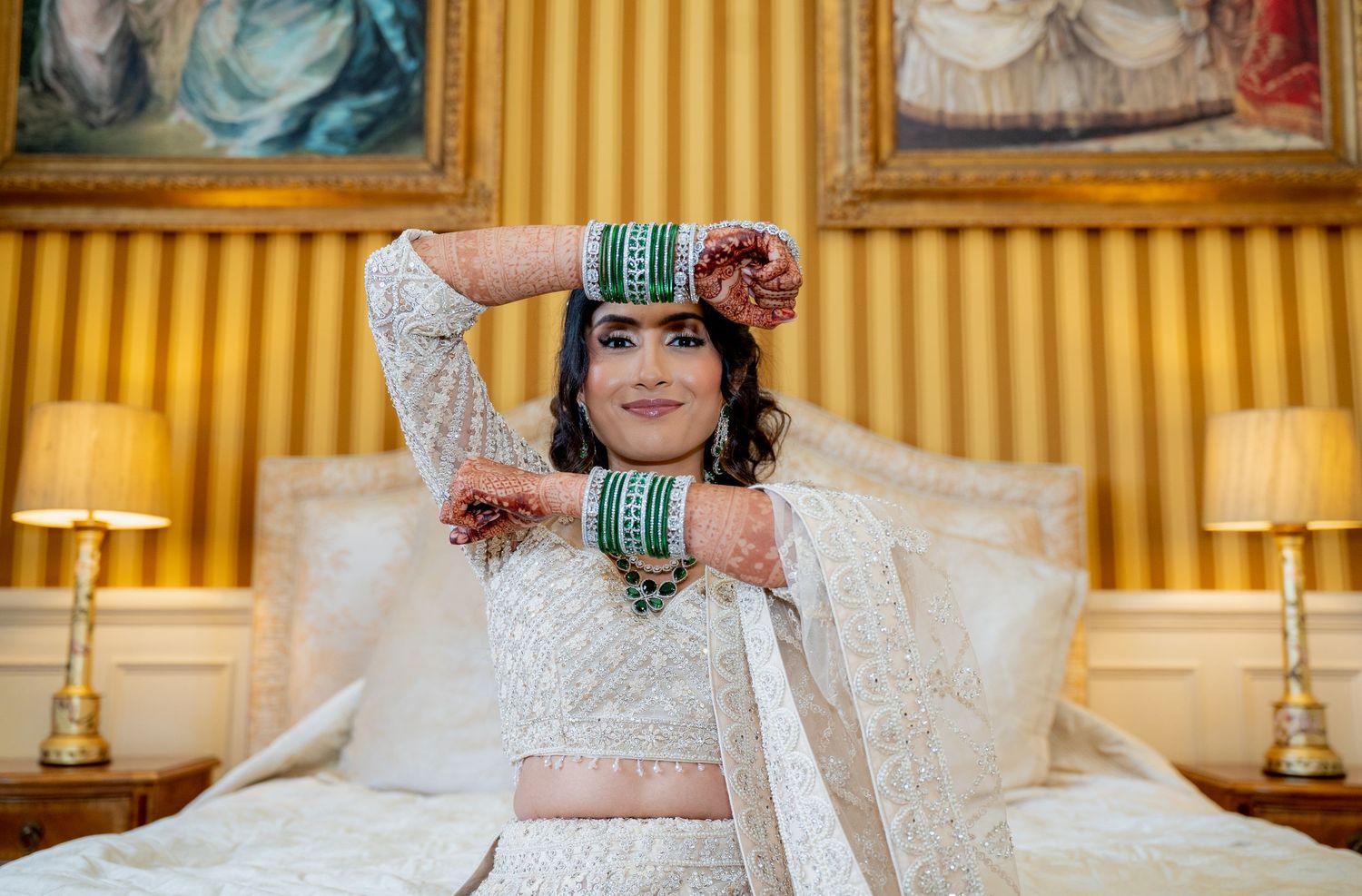 Indian bride in white lehenga with green bangles and henna sits elegantly on bed in golden room.