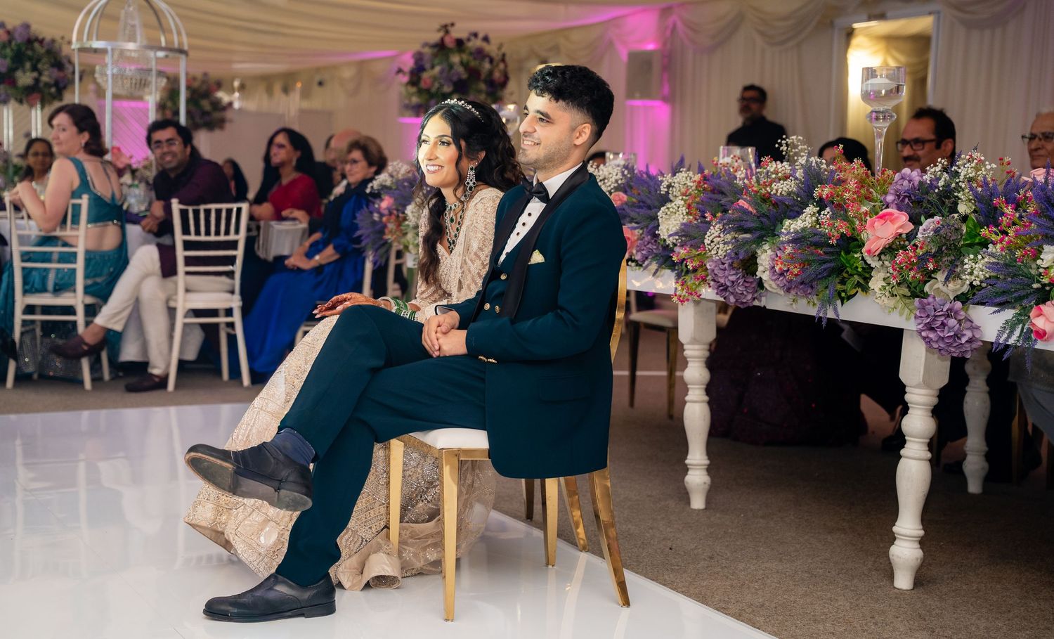 Bride and groom sitting together at wedding reception with colorful floral arrangements and guests in background.