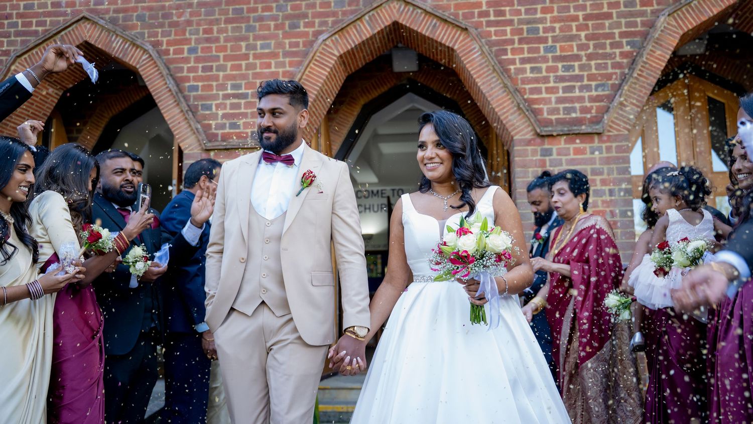 Groom in tan suit and bride in white gown exit church amid confetti, guests in festive attire surround them.