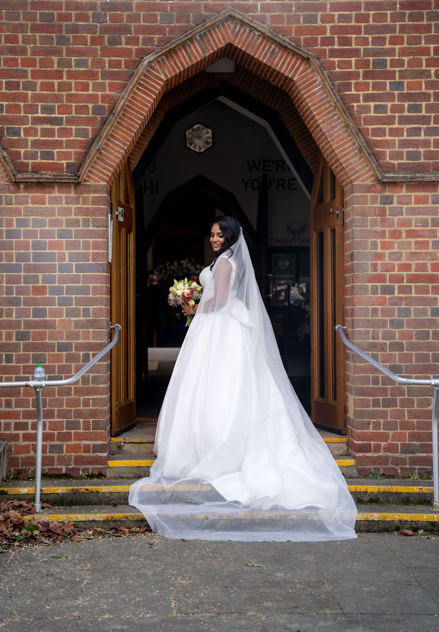 Bride in white gown and veil stands at church entrance holding bouquet, long train flowing on steps.