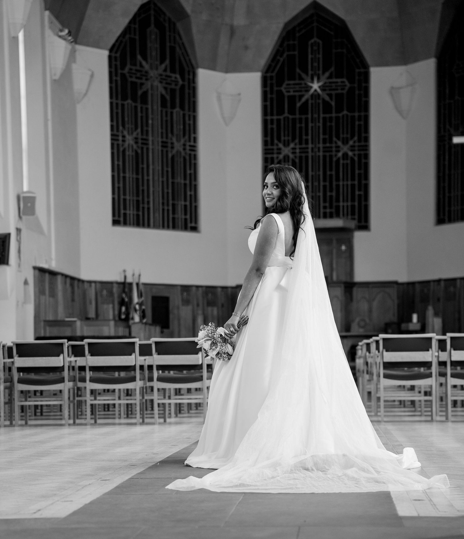 Black and white portrait of bride in elegant gown with long veil, holding bouquet inside a church.