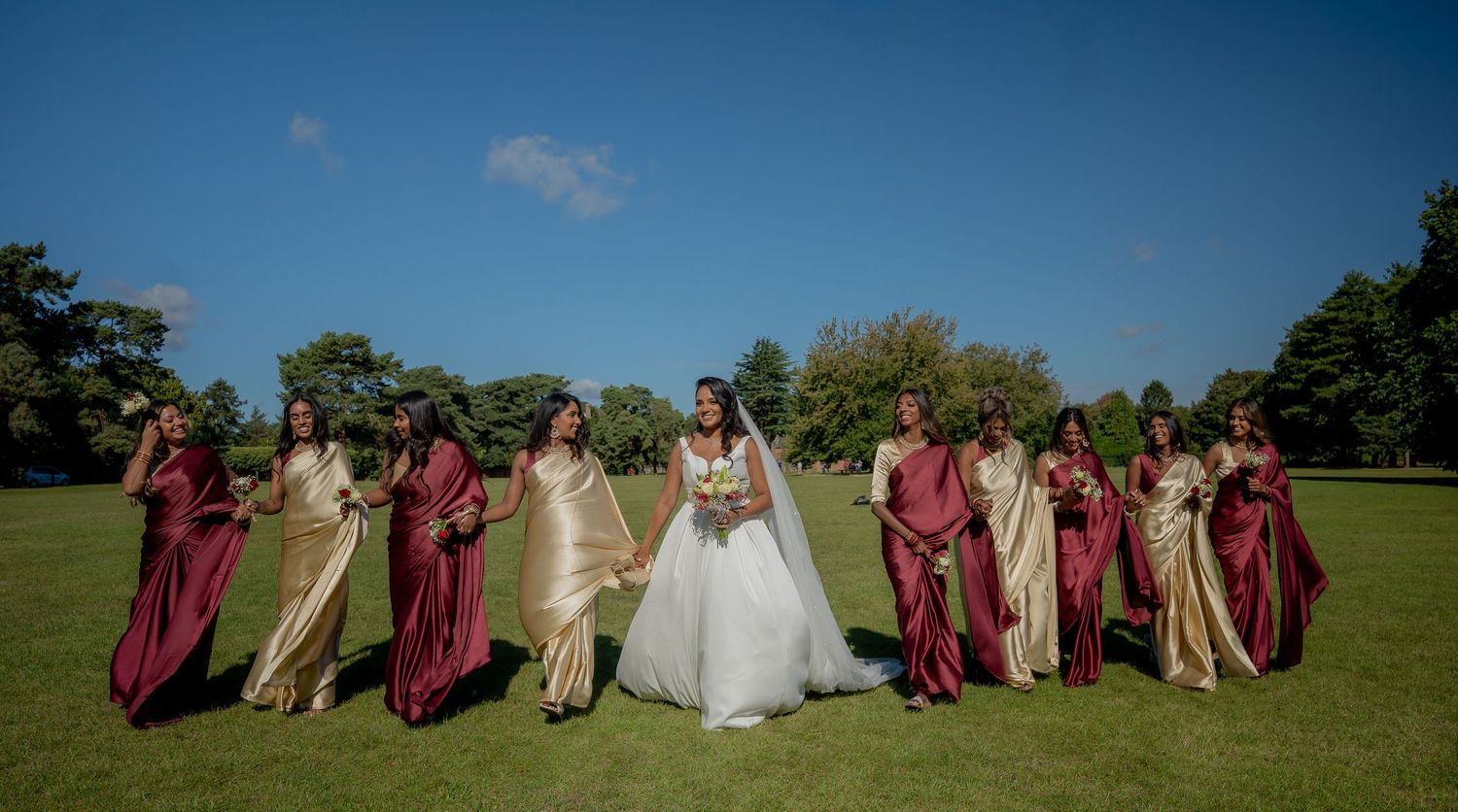 Bride in white ball gown flanked by bridesmaids in burgundy and gold dresses on sunny lawn.