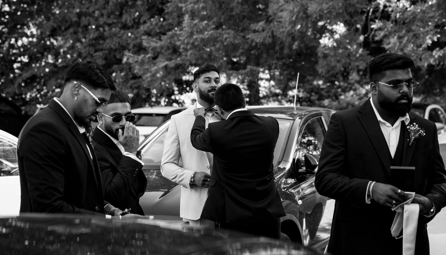 Group of men in suits embrace and greet each other near cars at an outdoor wedding in black and white.