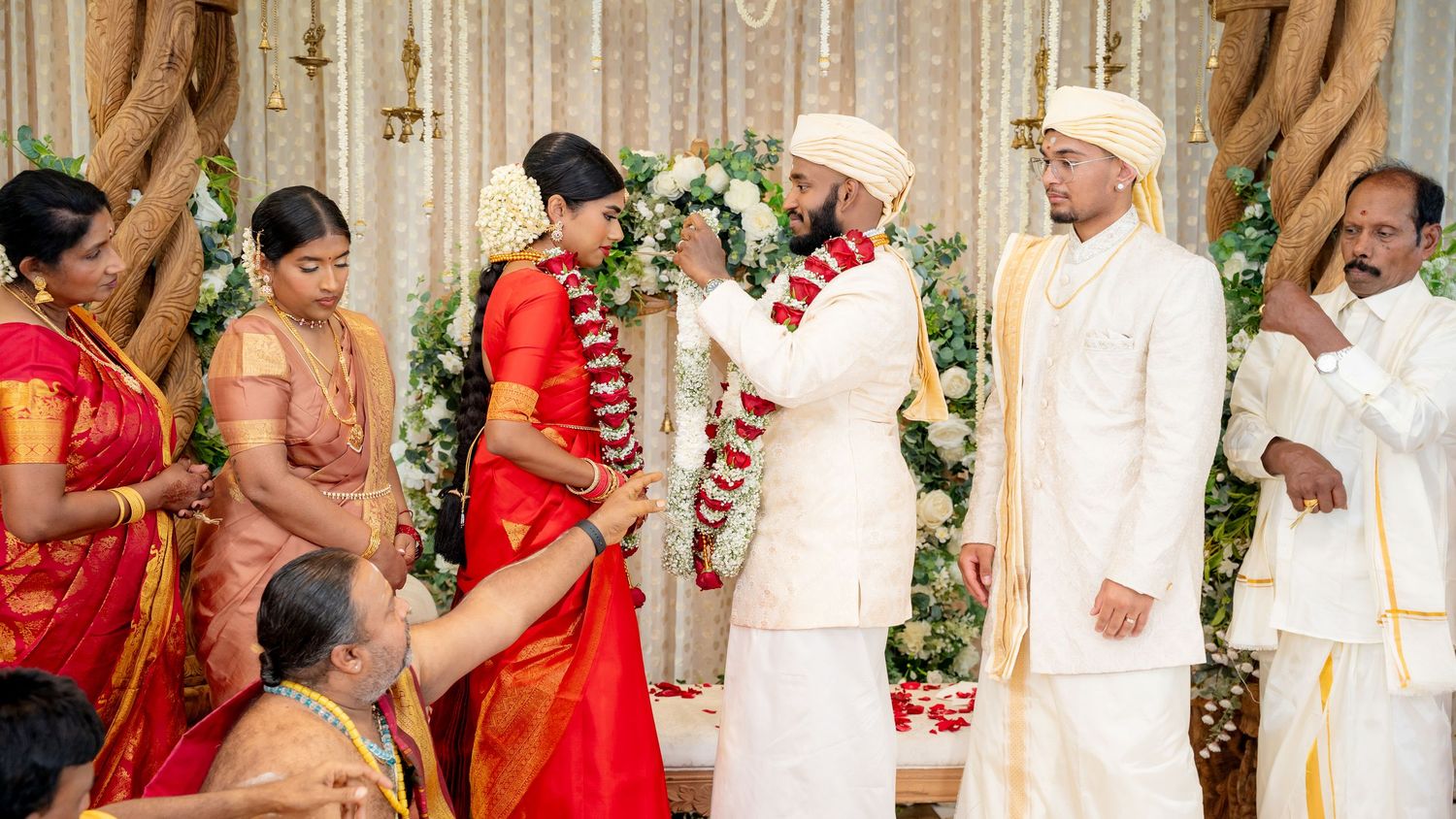 South Indian wedding couple exchanges floral garlands on decorated stage surrounded by family members.