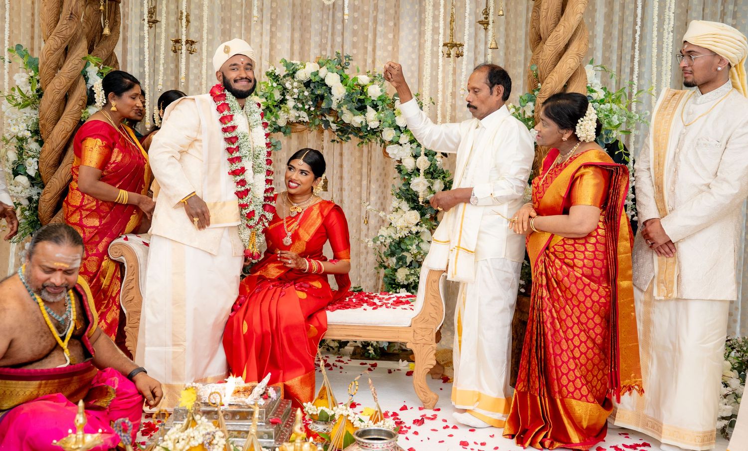 Groom in white garland stands with bride in red saree during South Indian Hindu wedding celebration.