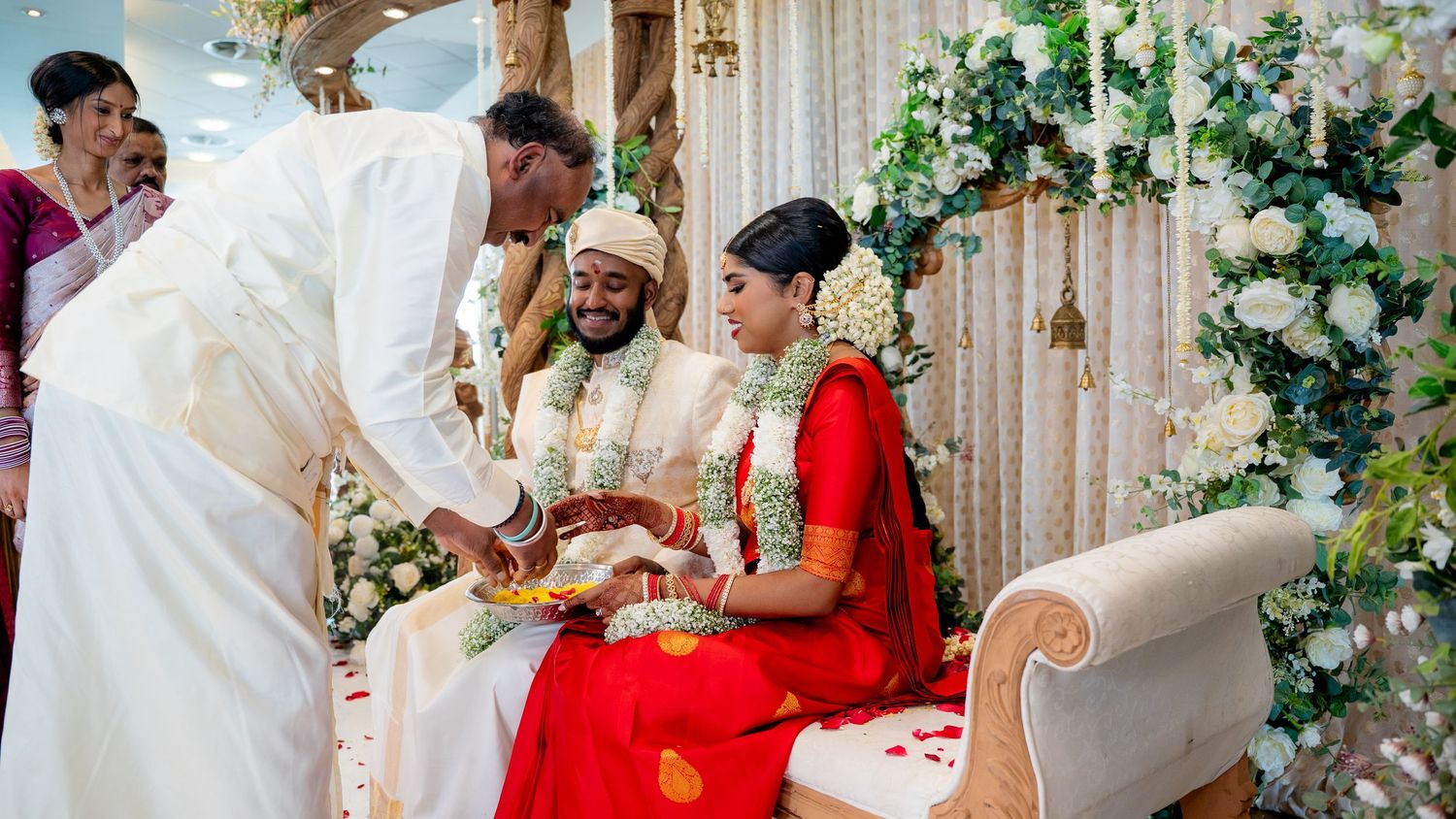 Elder man blessing Hindu bride and groom seated at beautifully decorated Indian wedding ceremony mandap.