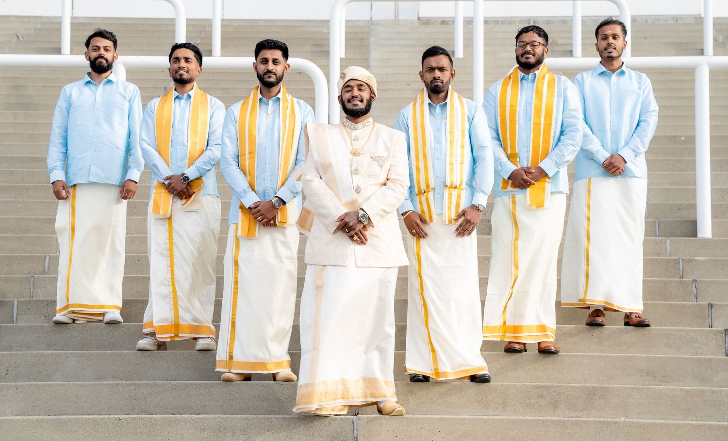 Group of seven men in traditional Kerala attire with white dhotis and gold borders posing on steps.