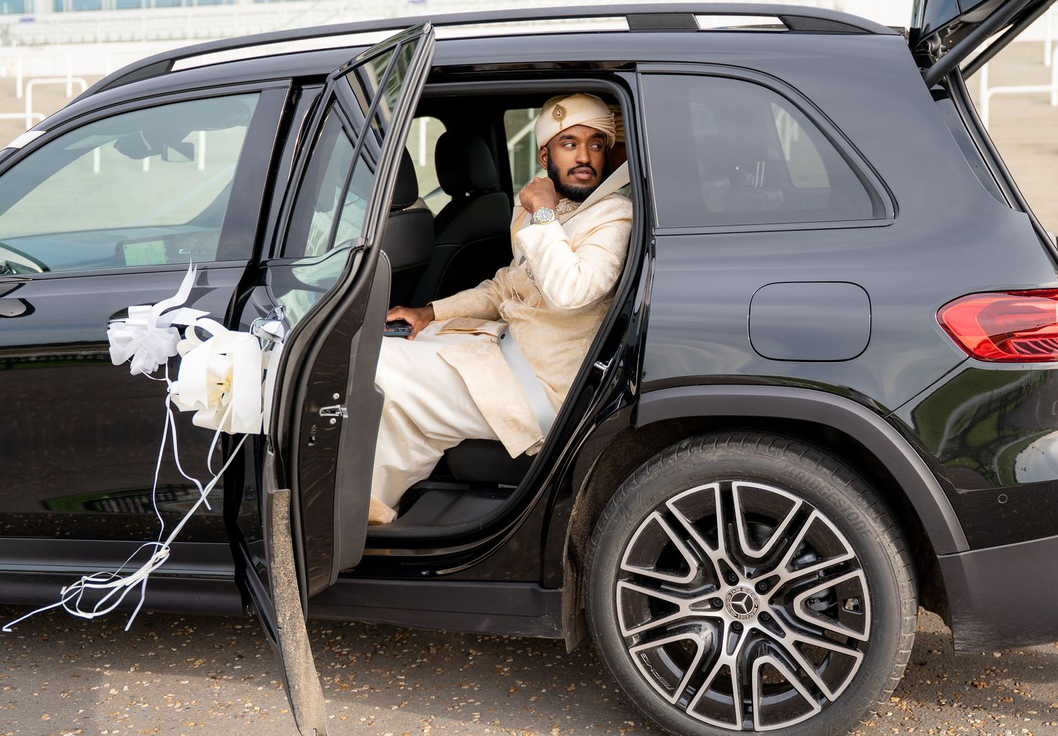 Man in white traditional attire sitting in a decorated black Mercedes SUV at an outdoor event.