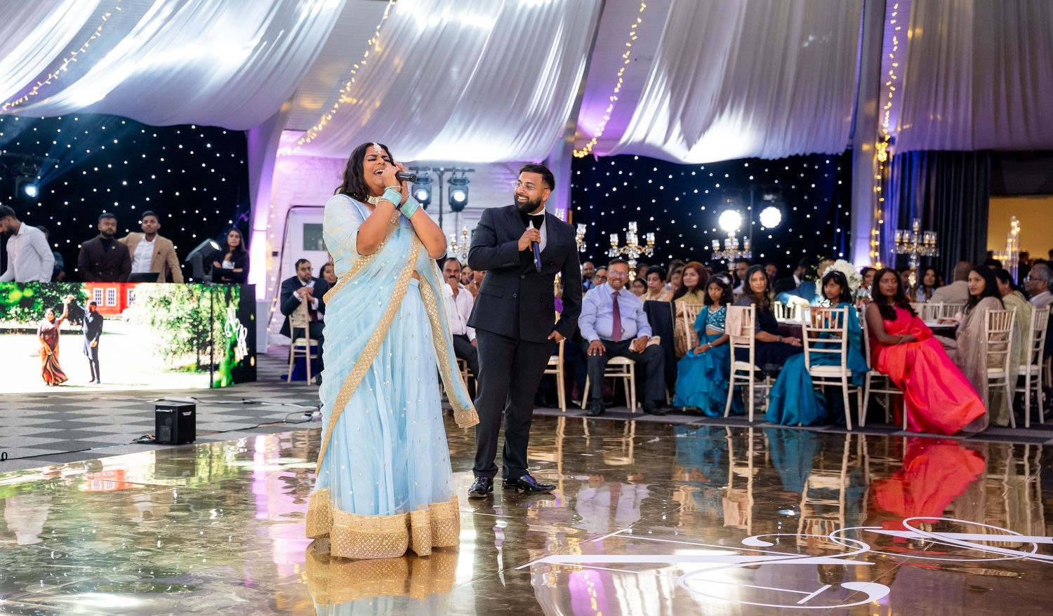 Couple in elegant attire performing at an Indian wedding reception in a decorated banquet hall with guests seated.