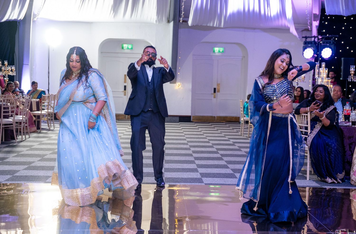 Three guests performing an energetic dance in colorful Indian outfits at a wedding reception.