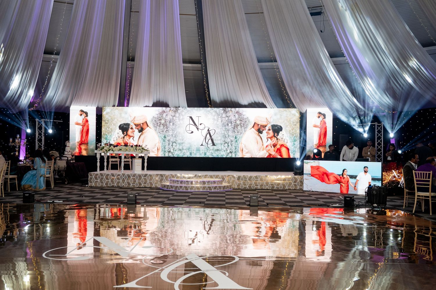 Elegant wedding reception venue with draped white ceiling, illuminated stage backdrop, and reflective dance floor.