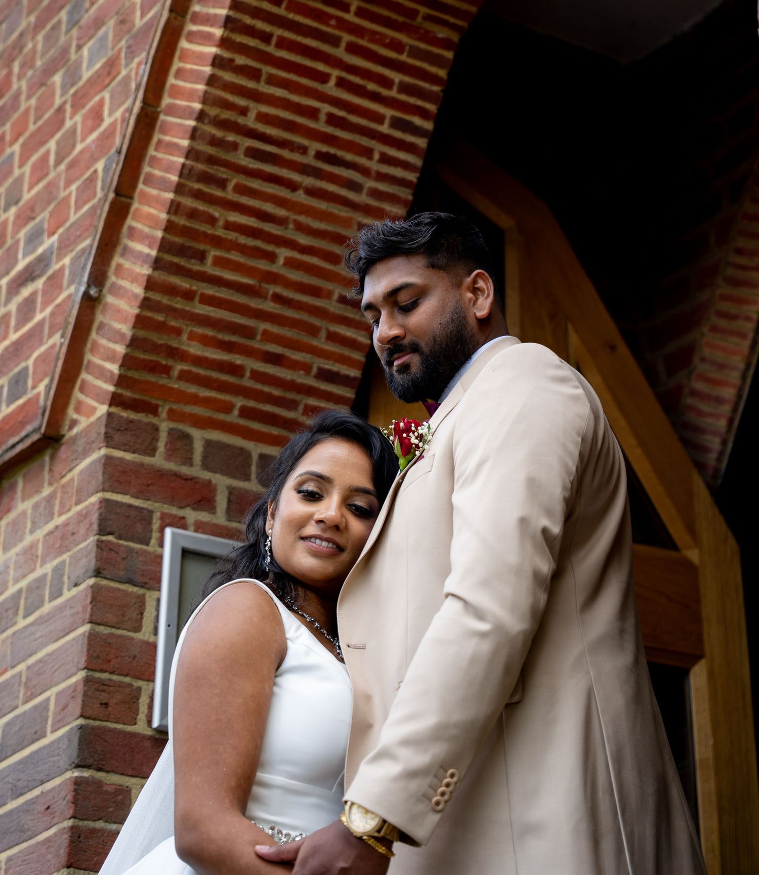 Couple in wedding attire poses by brick building, bride in white dress, groom in beige suit with floral boutonniere.