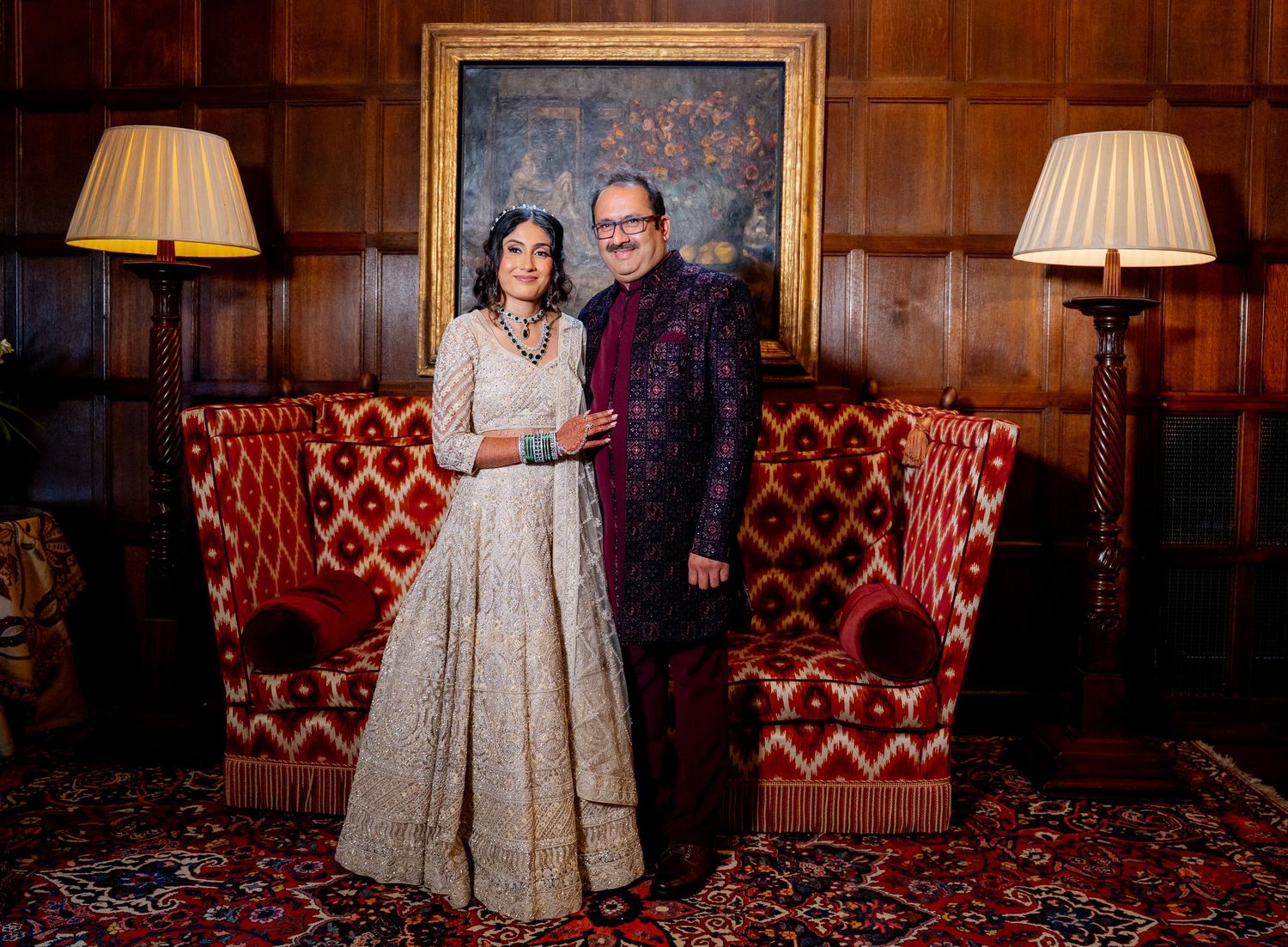 Couple in elegant South Asian attire posing in a wood-paneled room with red velvet chairs and warm lamp lighting.
