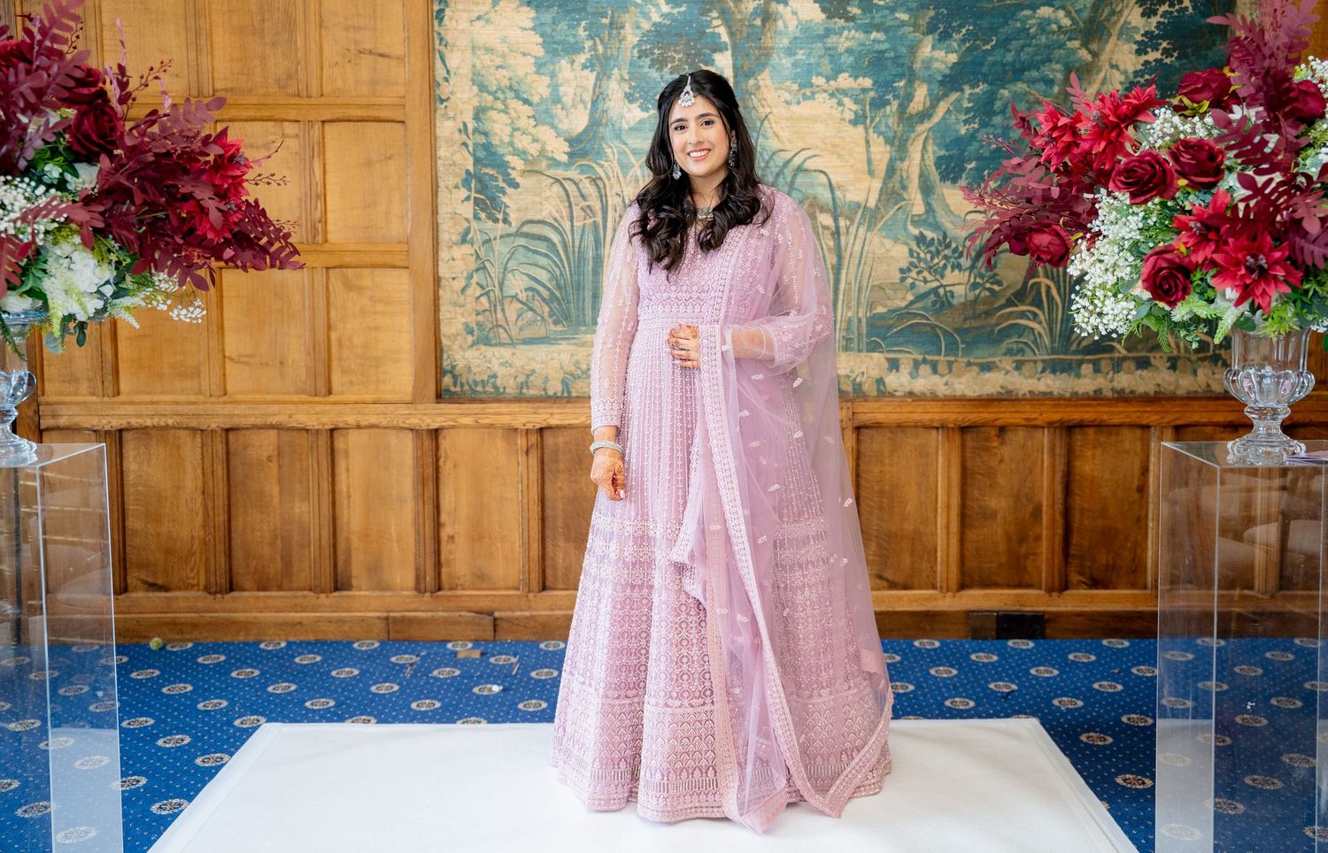 Woman in pink embroidered anarkali suit standing elegantly in a decorated room with floral arrangements.