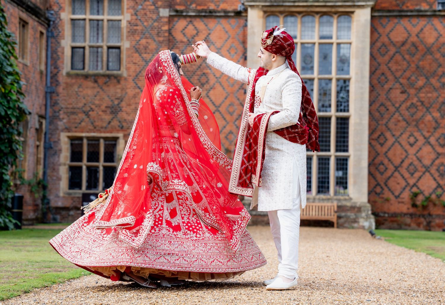 Indian groom spinning bride in stunning red embroidered lehenga during outdoor wedding photoshoot at historic manor.