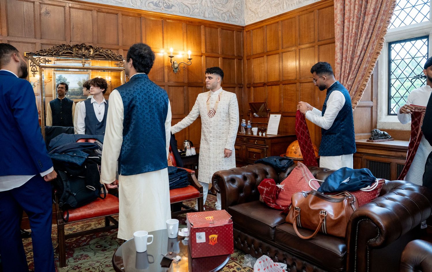 Indian groom in white sherwani with groomsmen in navy kurtas preparing in traditional wood-paneled room.