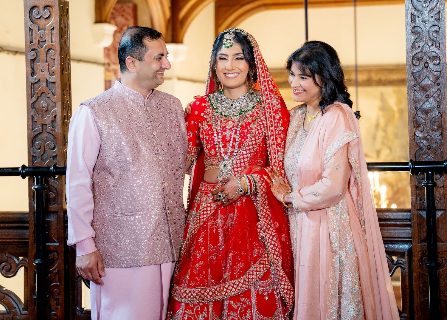 Indian bride in red lehenga posing with parents in ornate wedding venue.