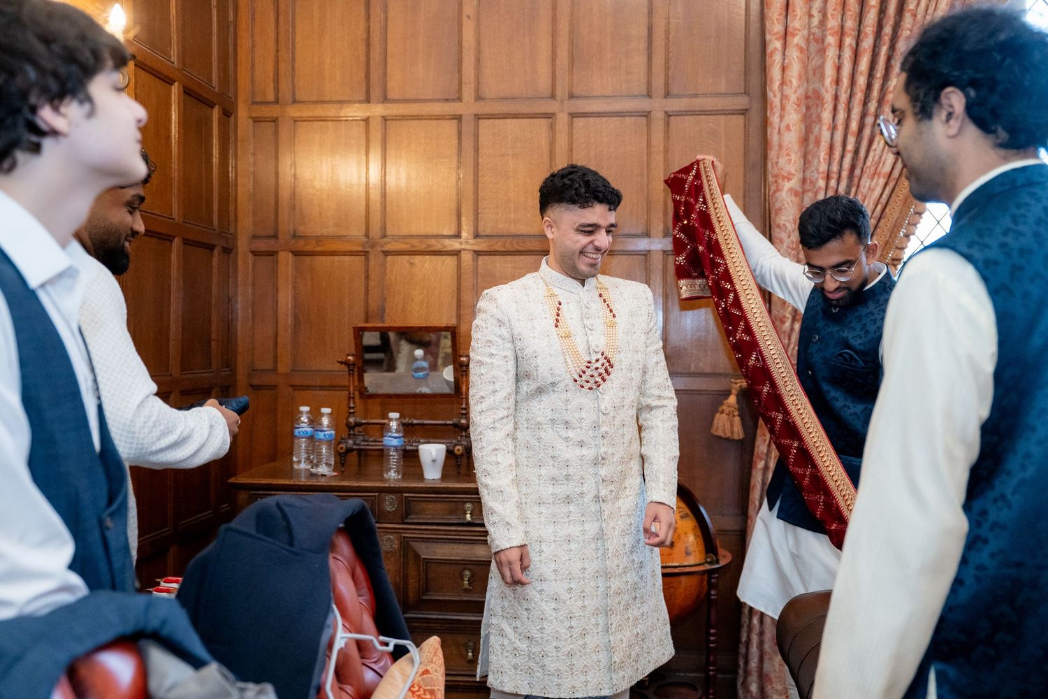 Groom in white sherwani smiling as groomsmen help prepare red wedding dupatta in wood-paneled room.