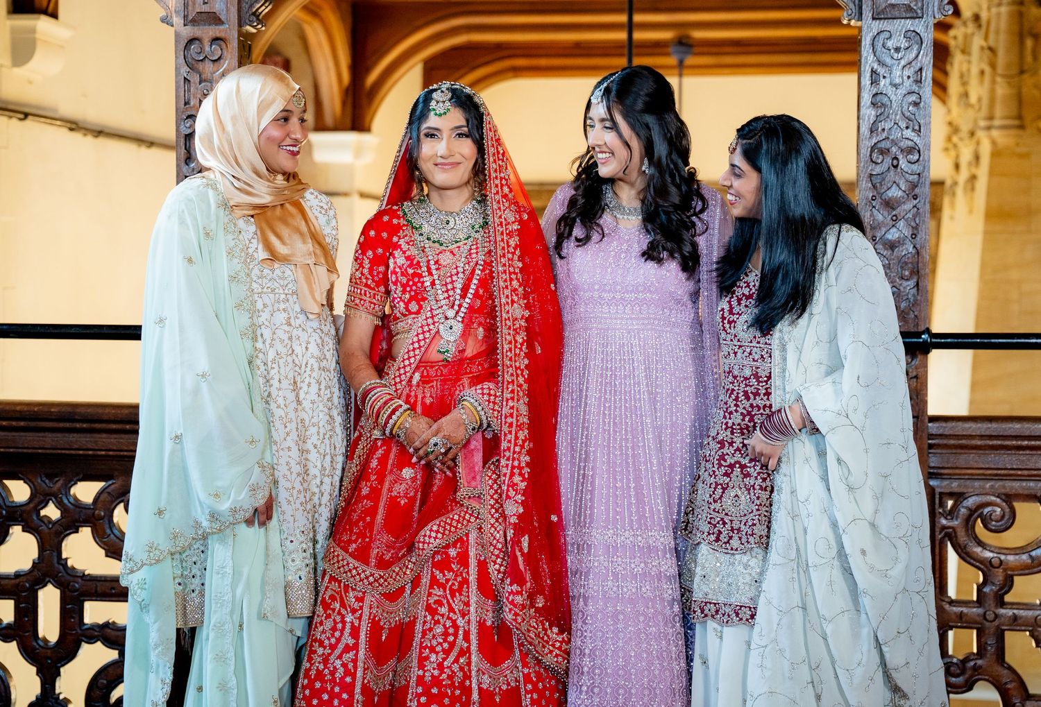 Indian bride in red lehenga poses joyfully with three friends in pastel outfits.