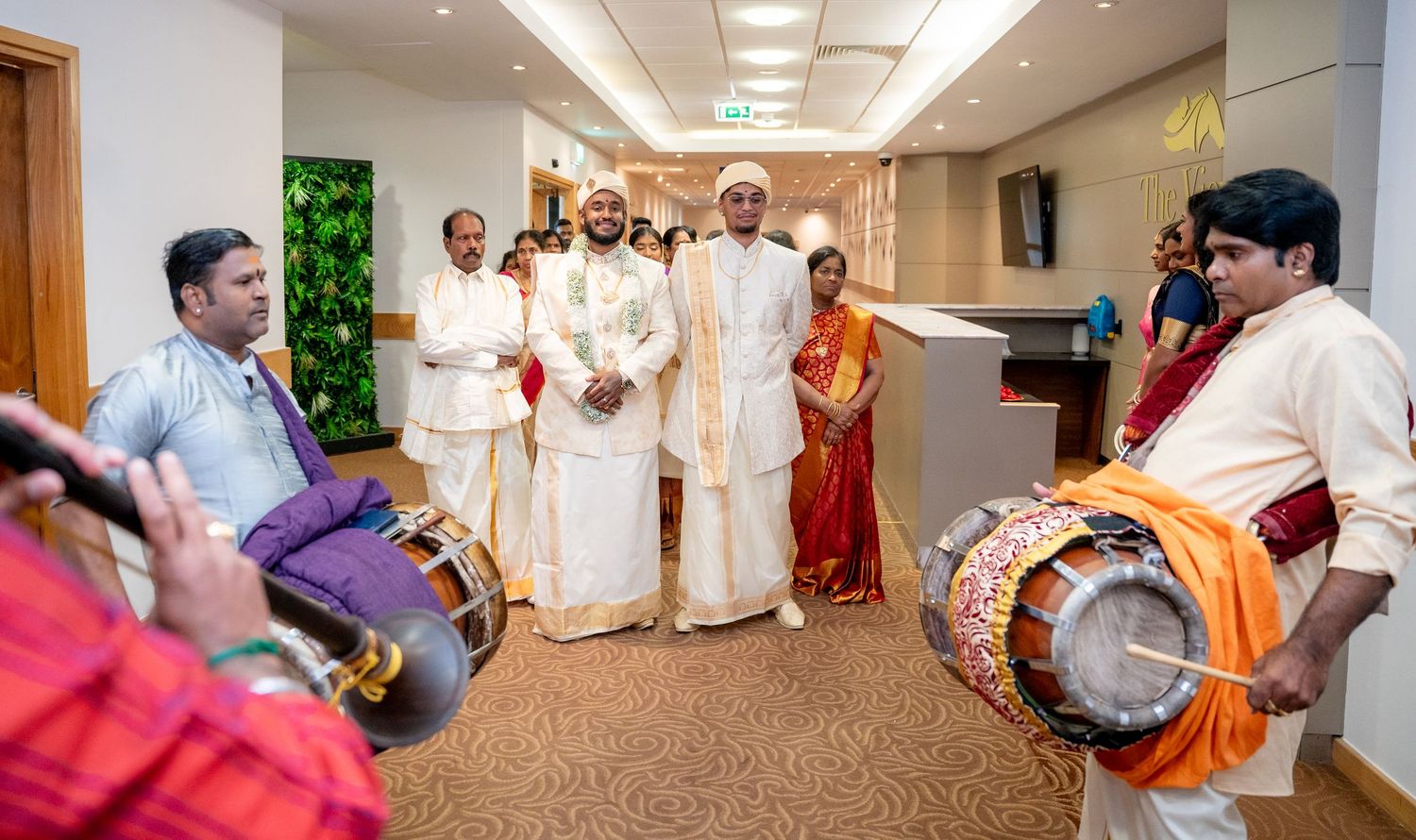 Traditional South Indian wedding ceremony with drummers playing chenda, guests in white mundu and saree attire in elegant venue.