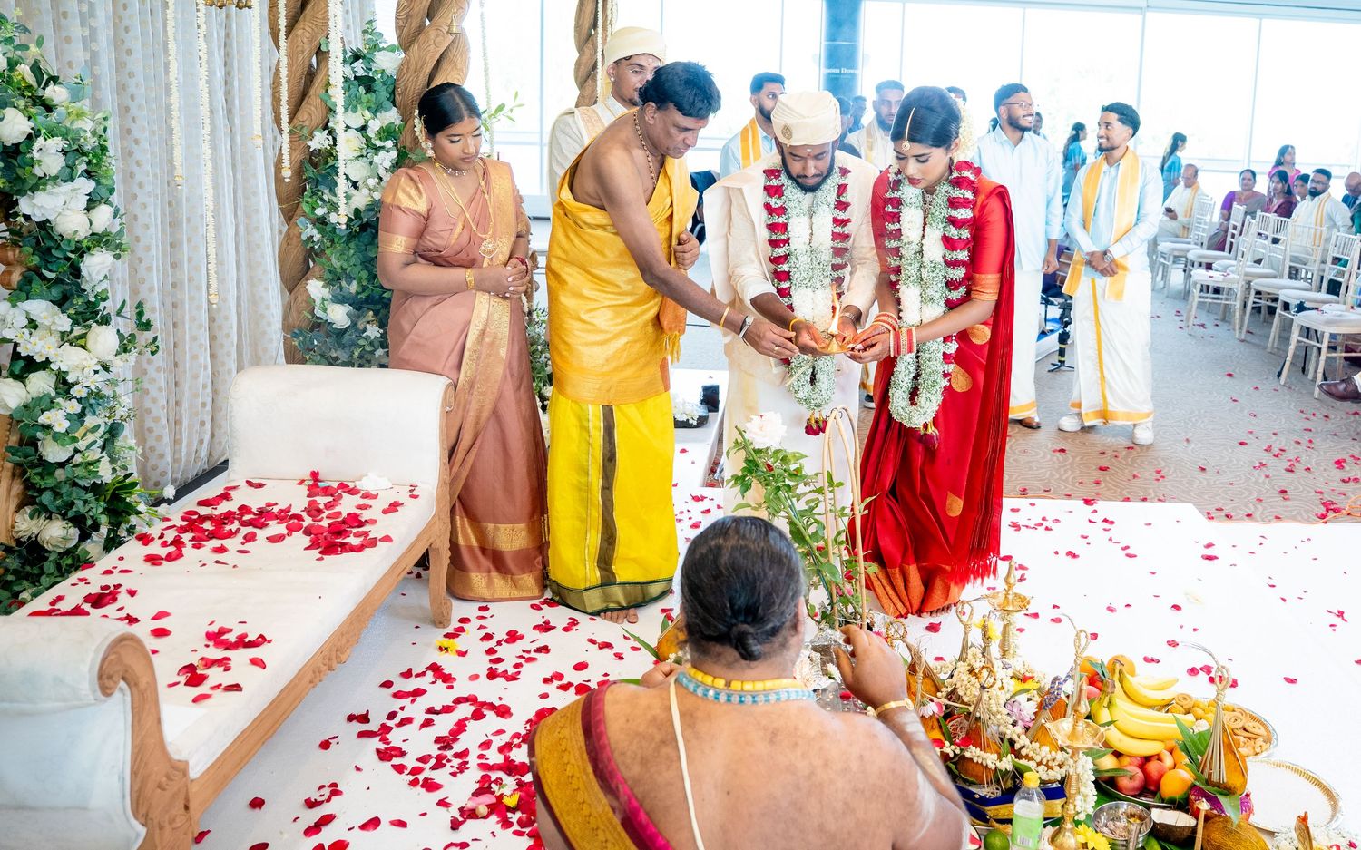 Priest performs sacred Hindu wedding rituals for couple surrounded by family and flower petals.