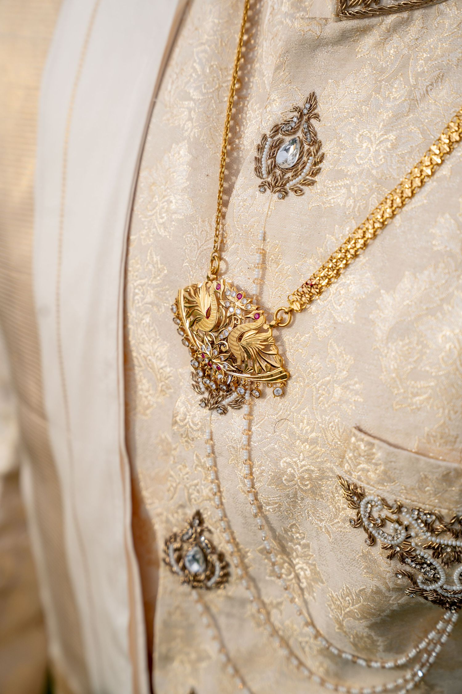 Close-up of ornate gold jewelry on cream brocade Indian wedding sherwani with intricate embroidery and gemstone details.