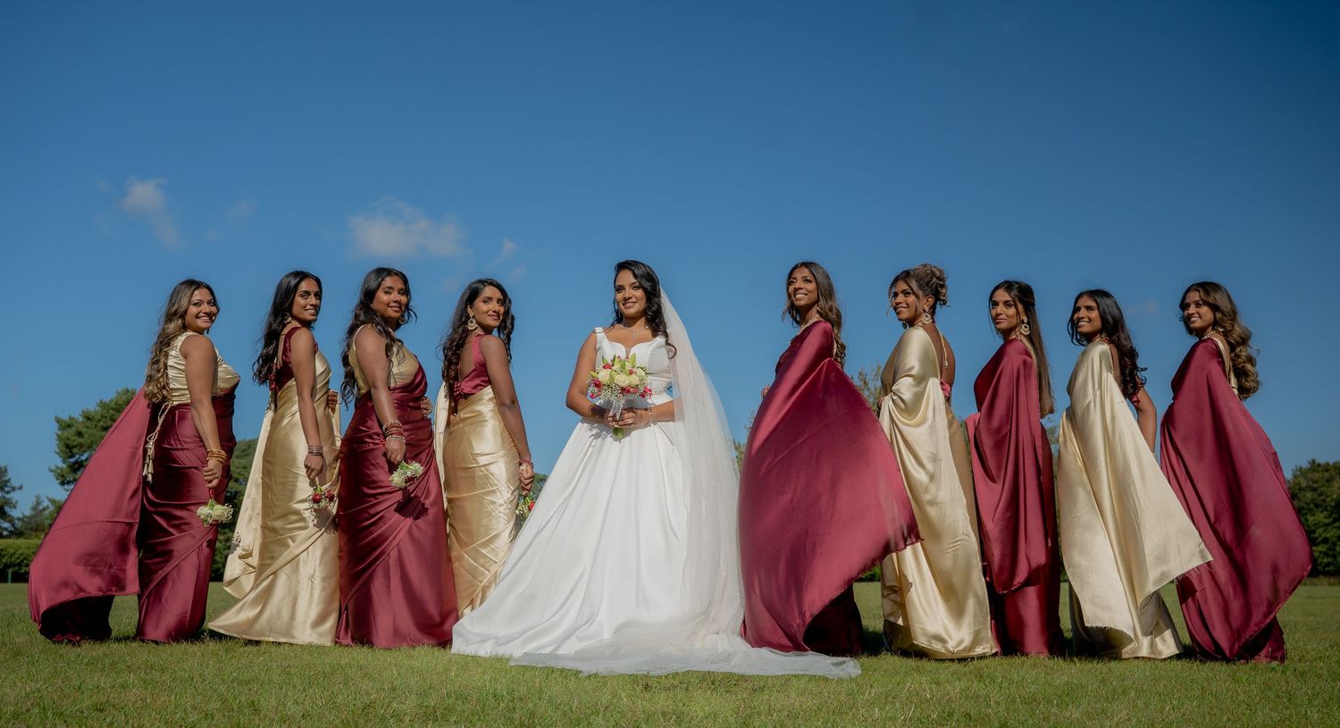Bride in white gown poses with ten bridesmaids in burgundy and gold dresses under blue sky.