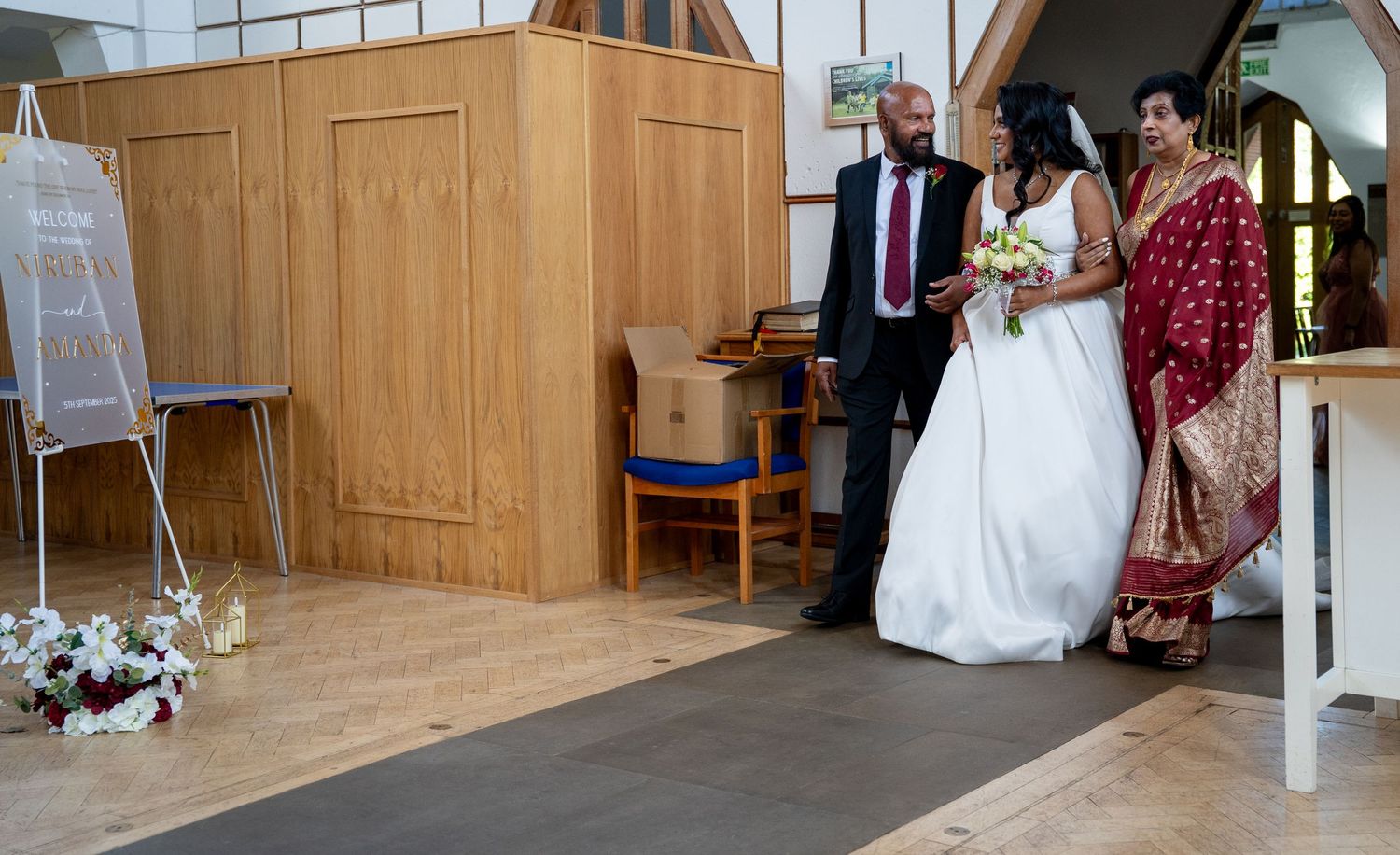 Bride in white gown walks down church aisle escorted by parents in formal and traditional Indian attire.