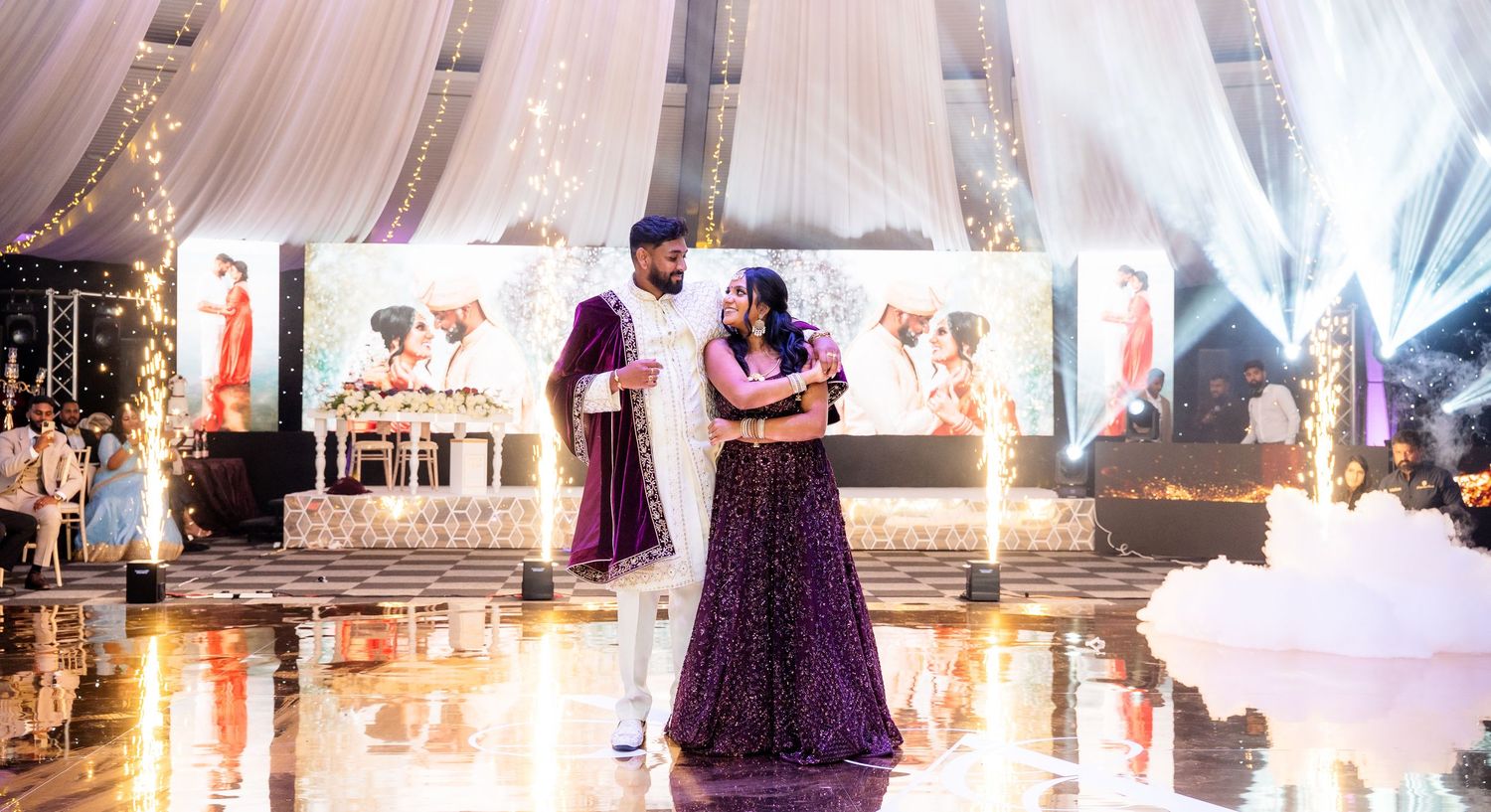 Couple shares first dance at elegant Indian wedding reception with sparkling stage, purple gown, and white sherwani.