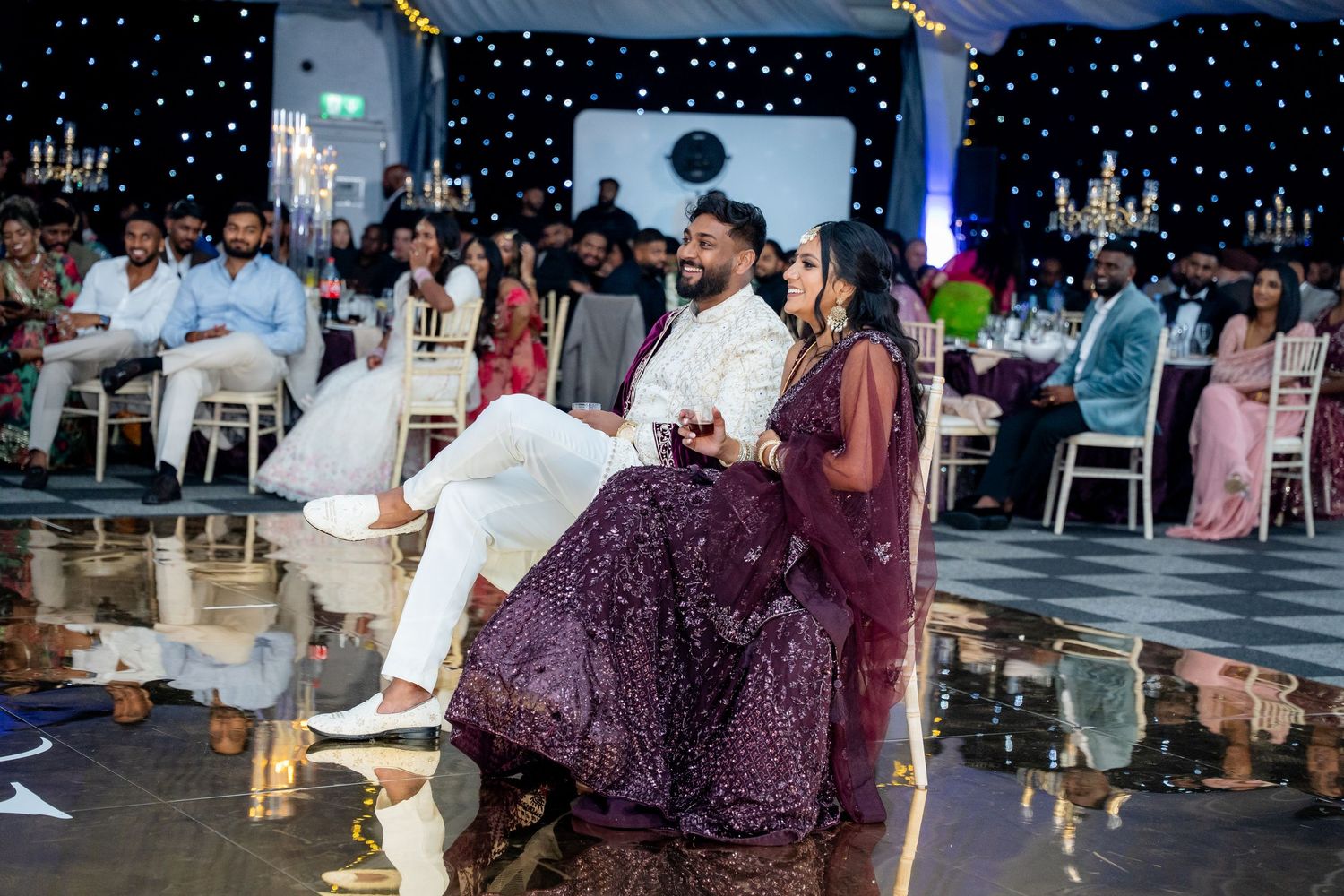 Bride in purple lehenga and groom in white sherwani sit together on the wedding reception dance floor.