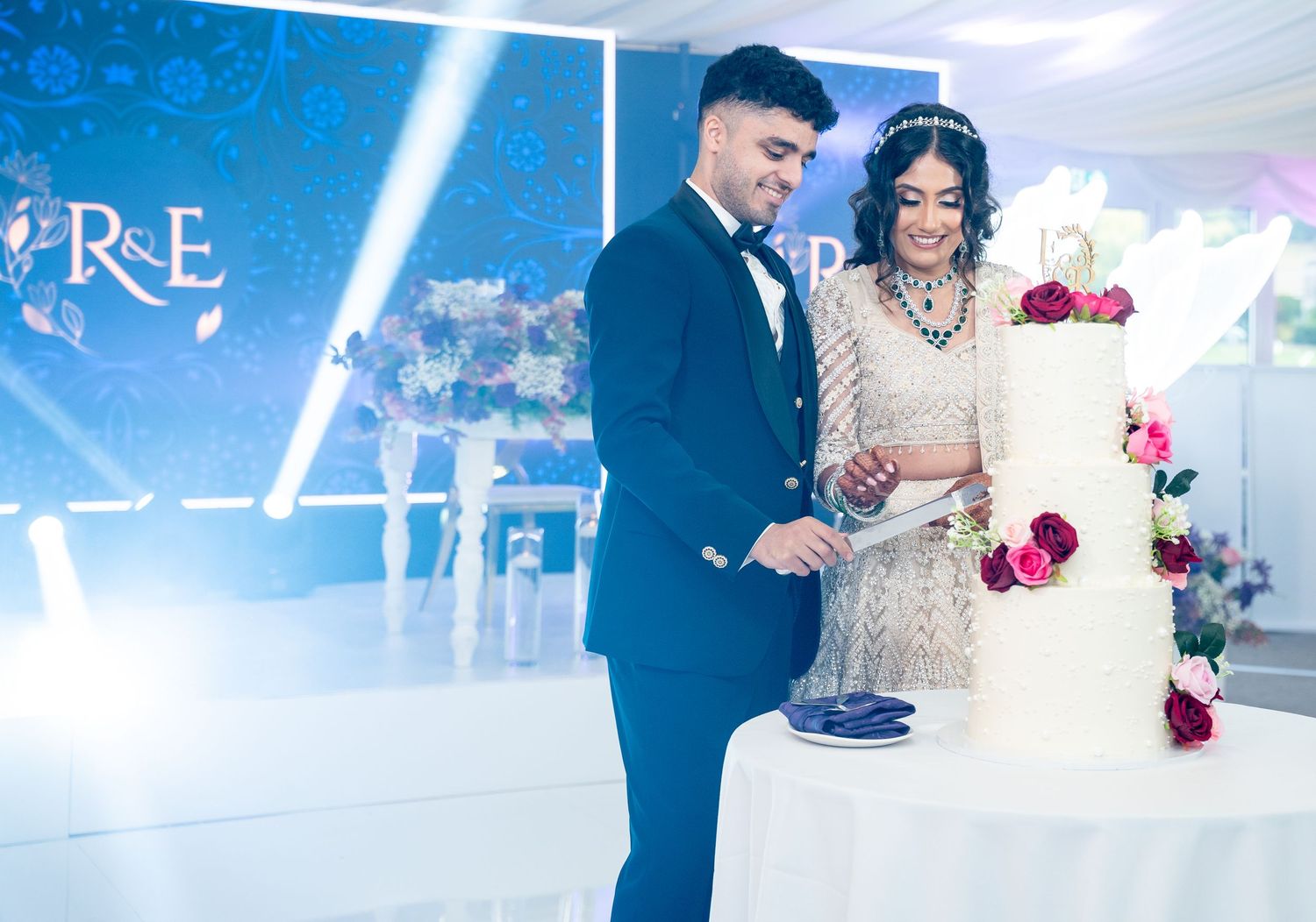 Couple cutting elegant white three-tier wedding cake with R&E monogram backdrop at wedding reception.