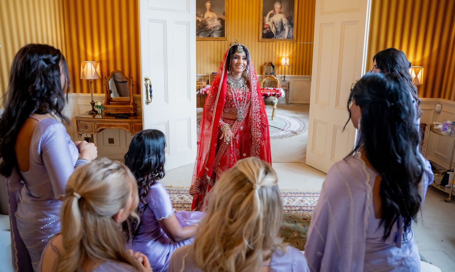 Indian bride in red lehenga stands surrounded by bridesmaids in lavender outfits indoors.
