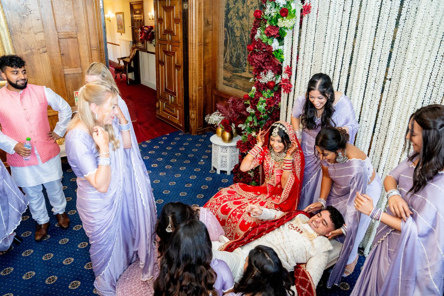 South Asian bride in red lehenga sits with groom during traditional wedding ceremony surrounded by bridesmaids in lavender.