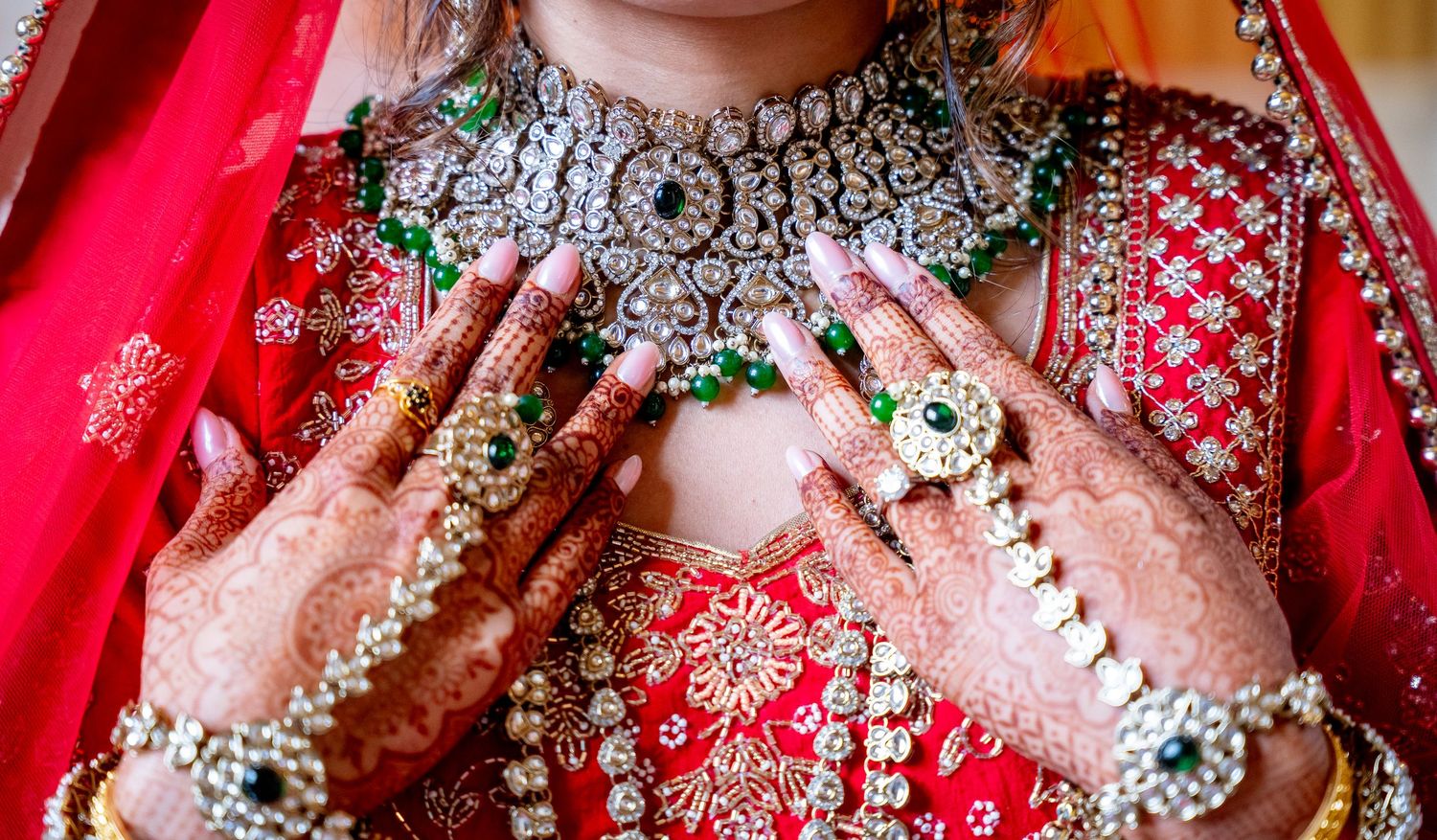 Indian bride showcasing intricate mehndi henna hands,diamond and emerald bridal jewelry,and red embroidered lehenga.