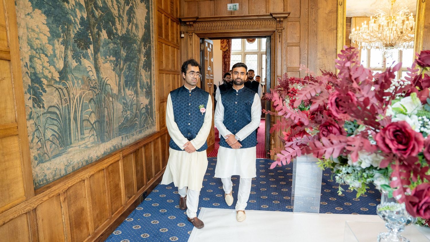 Two men in navy blue Nehru jackets walk through elegant wood-paneled hall at Indian wedding.