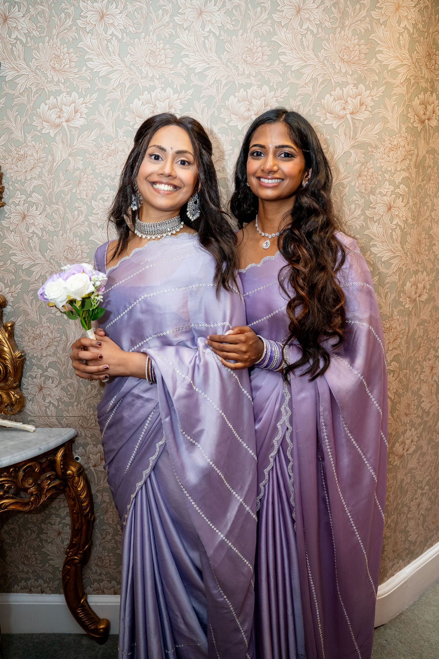 Two women in matching lavender sarees smile together, one holding white roses, at Indian wedding.