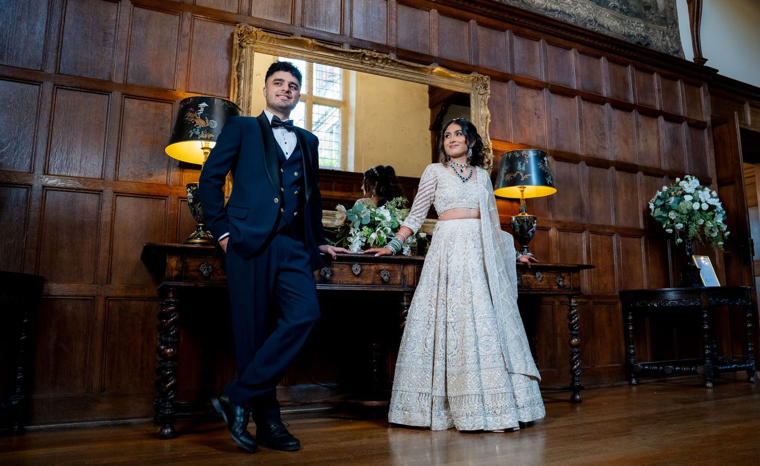 Elegant couple in wedding attire posing in a wood-paneled hall, groom in navy suit, bride in embroidered lehenga.