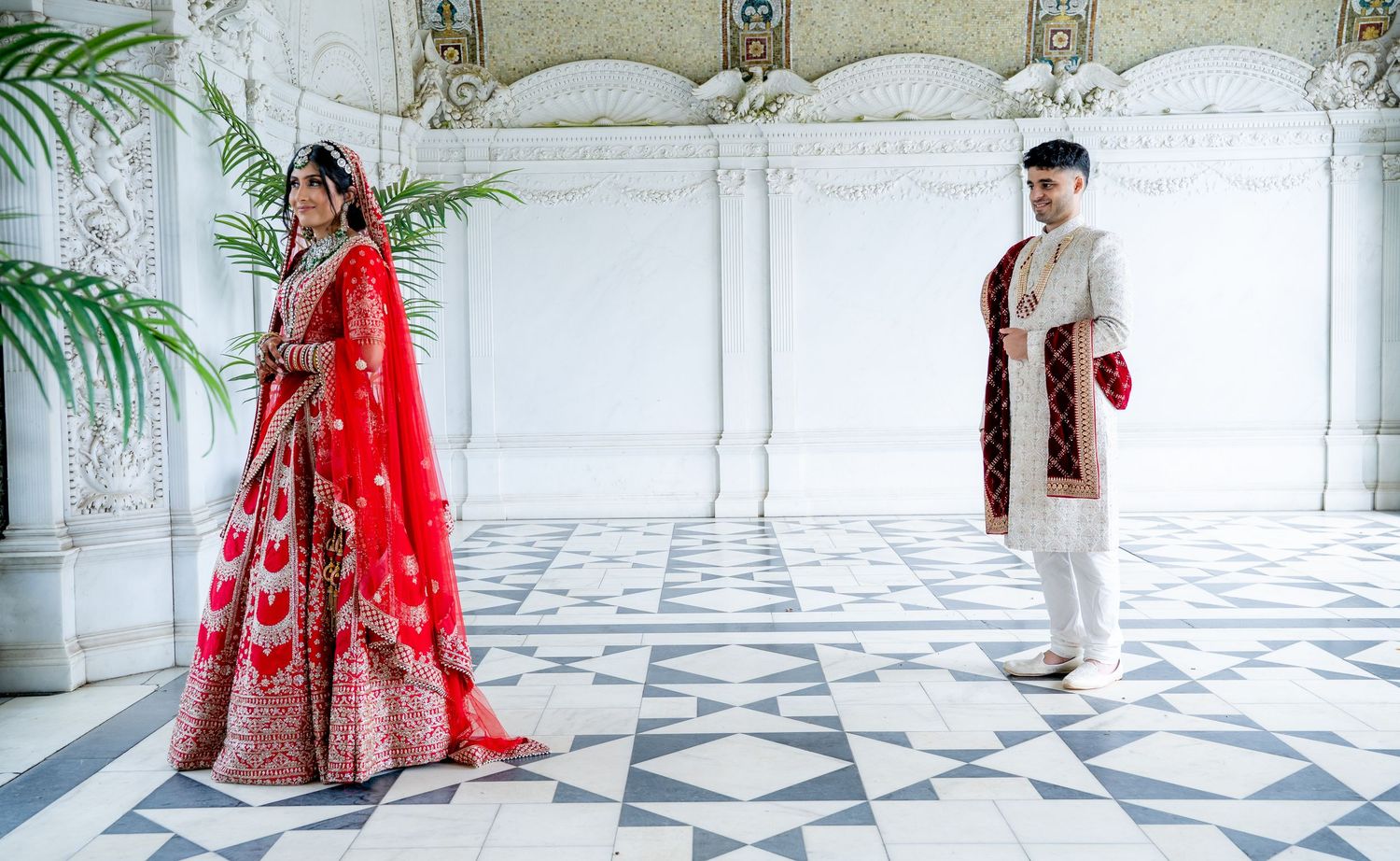 Indian bride in red lehenga and groom in white sherwani pose in a marble-floored venue.