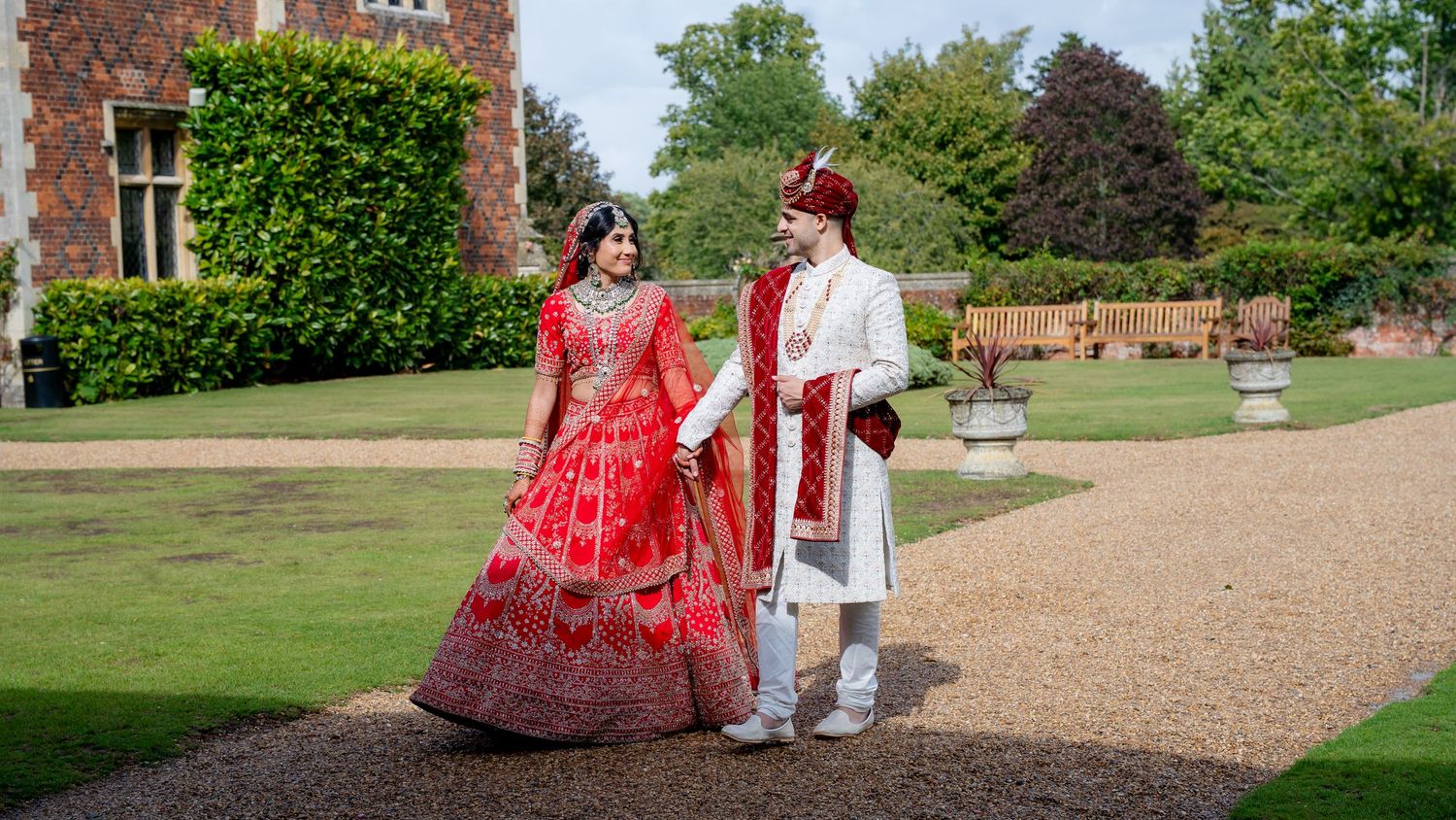Indian bride in red lehenga and groom in white sherwani walk hand in hand in a sunlit garden.